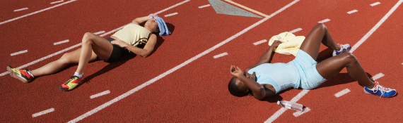 Two female student athletes laying on a track in the sun with towels and water bottles, battling the heat